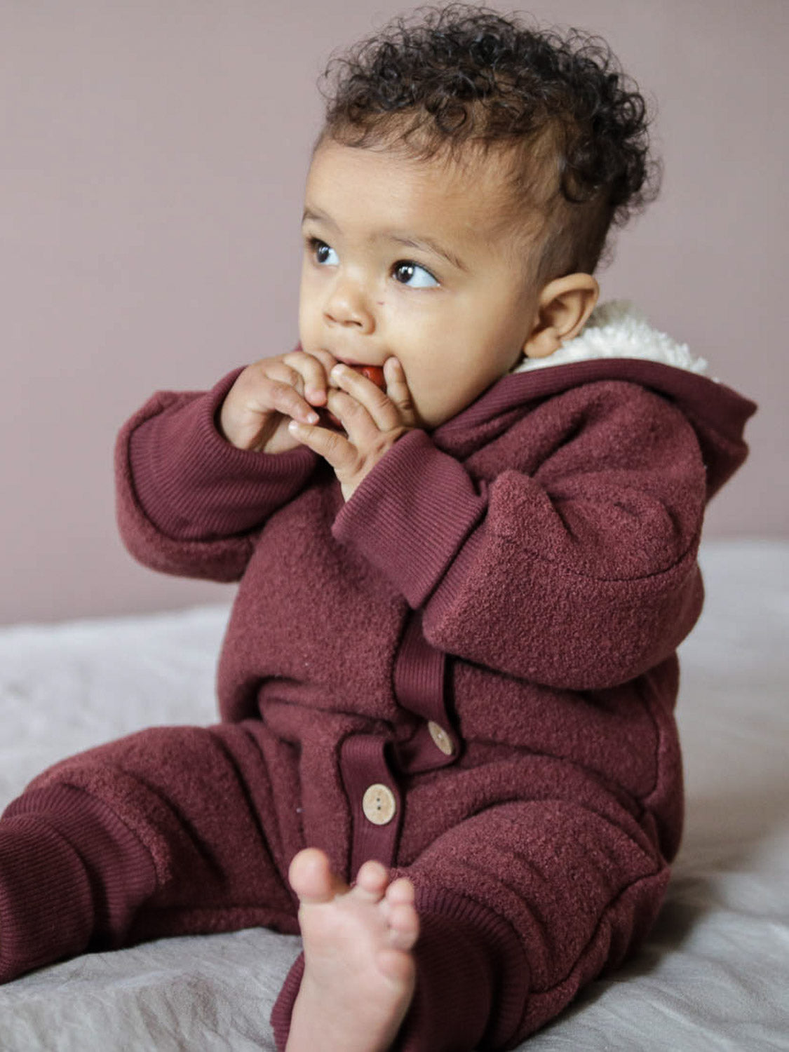 Baby in einem kuscheligen Teddy-Kuschelanzug Wollwalk - Kastanie mit Teddyfutter, ideal für kalte Tage, sitzt auf einem Bett und zeigt Details wie Krempelbündchen.