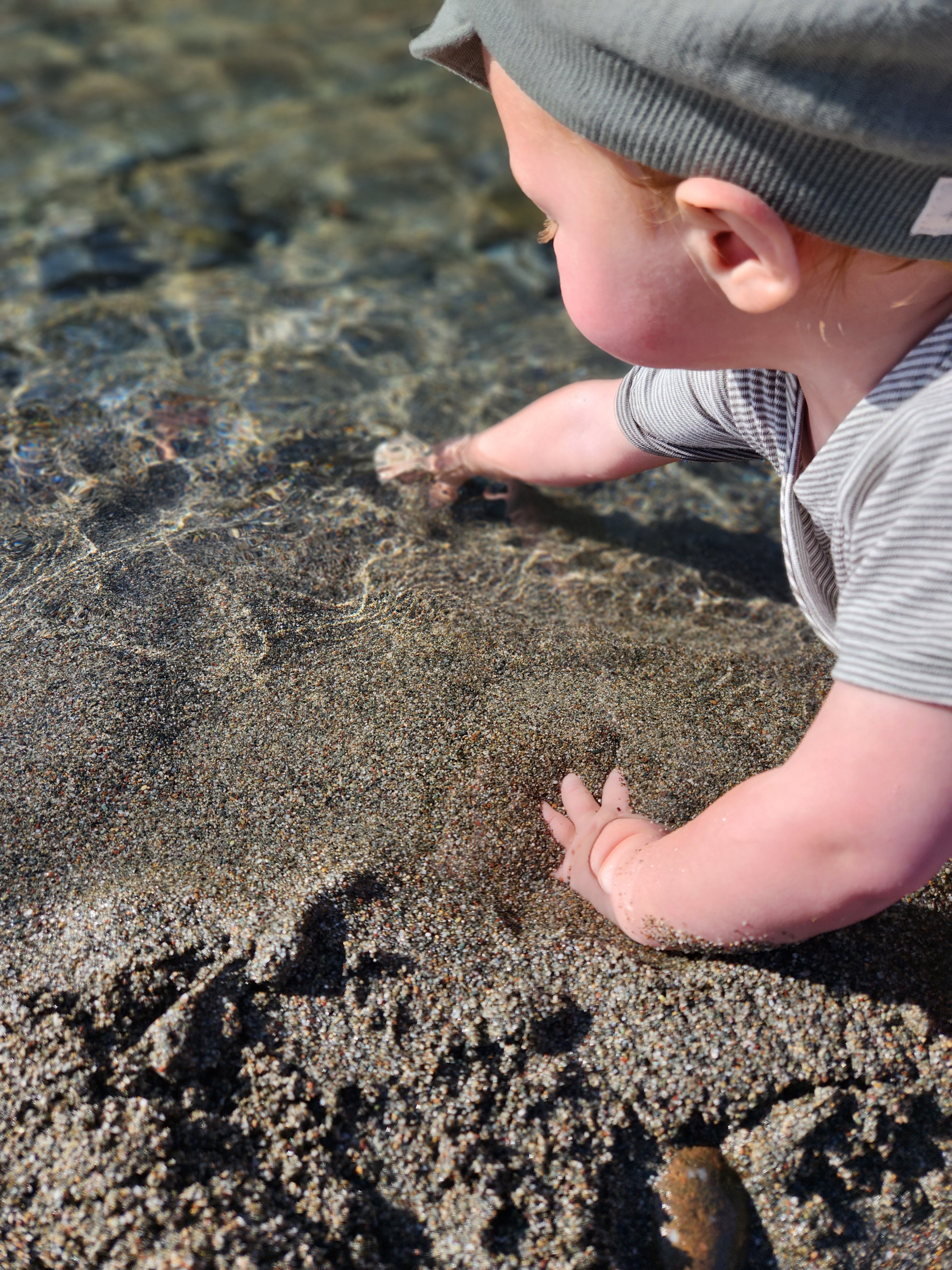 Ein Kind spielt am Strand und trägt die Michelmütze Bio-Musselin - Taube, eine luftige Sommermütze aus Bio-Baumwolle zum Schutz vor der Sonne.