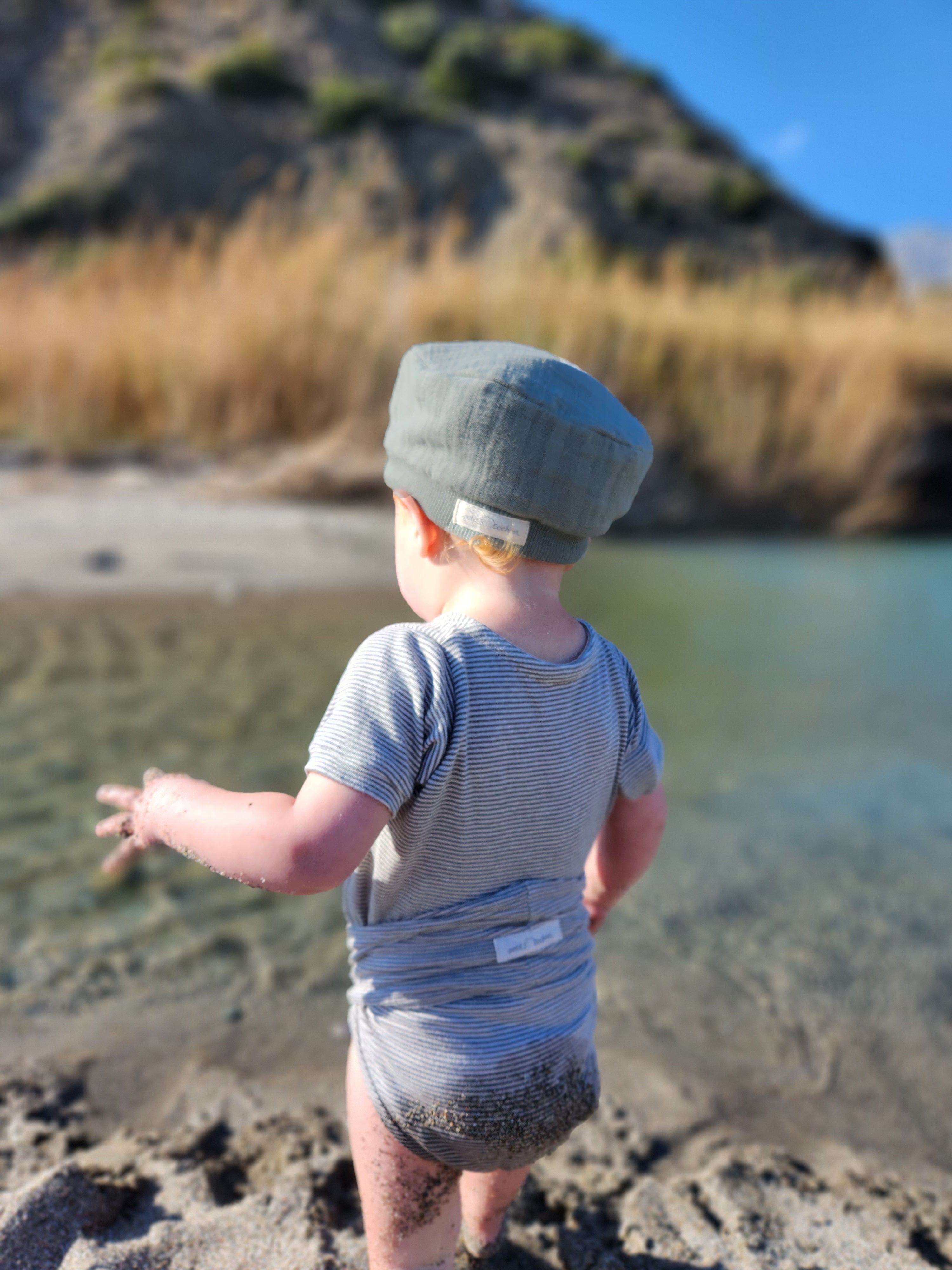 Kind am Strand trägt die Michelmütze Bio-Musselin - Taube, eine luftige Sommermütze aus Bio-Baumwolle mit elastischem Bündchen, die den Kopf vor Sonne schützt.