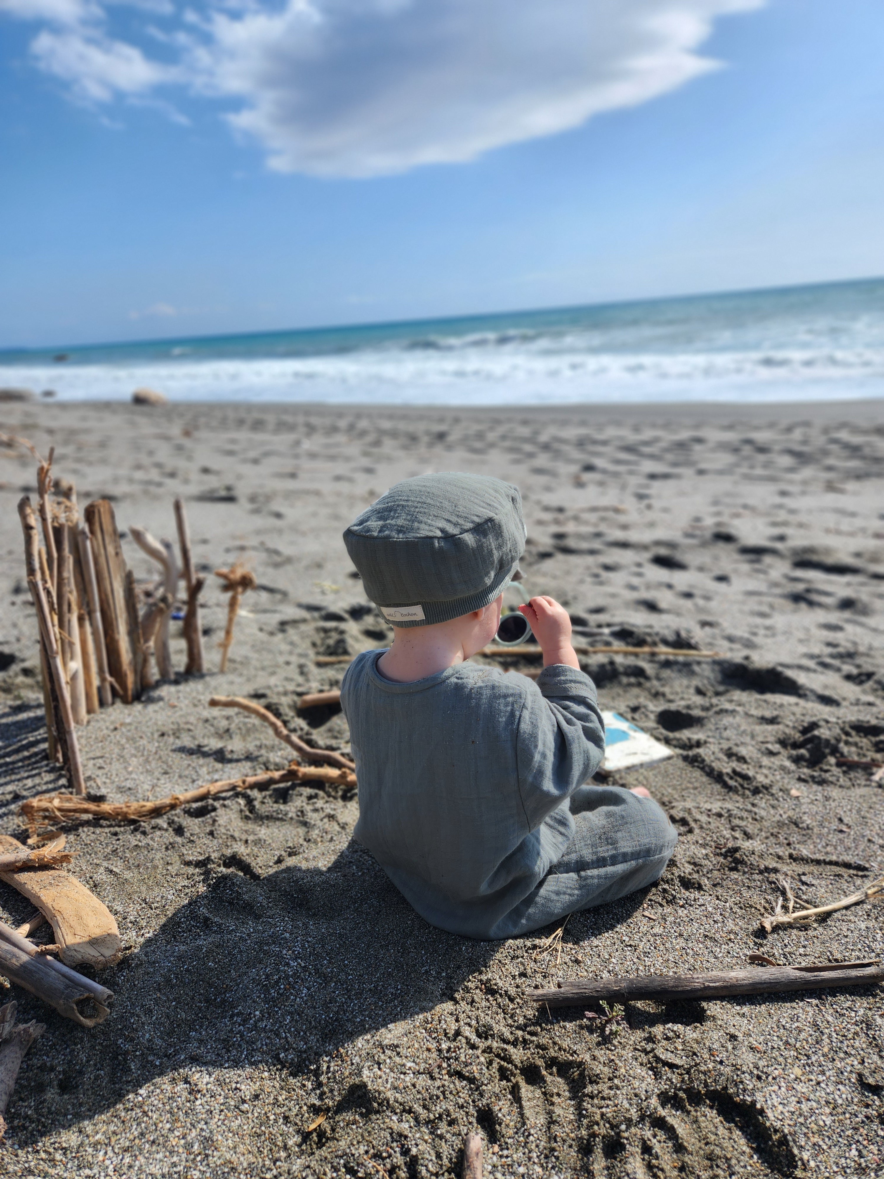 Kind auf Strand mit Michelmütze Bio-Musselin - Taube, sitzt am Meer. Die Mütze schützt vor Sonne, ist leicht und aus nachhaltigem Musselin gefertigt.